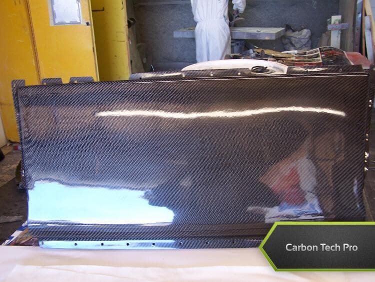 Precision-molded carbon fiber cover with custom mounting flanges. Large molded carbon fiber cover with mounting hole tabs along the edges standing upright.