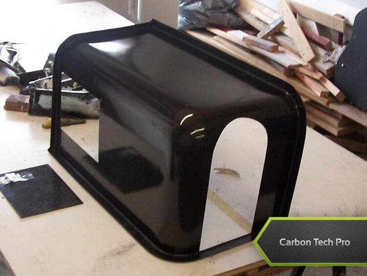 Smooth, high-gloss carbon composite housing built for custom aerospace applications. Glossy black molded housing component with cutouts resting upside down on a table.