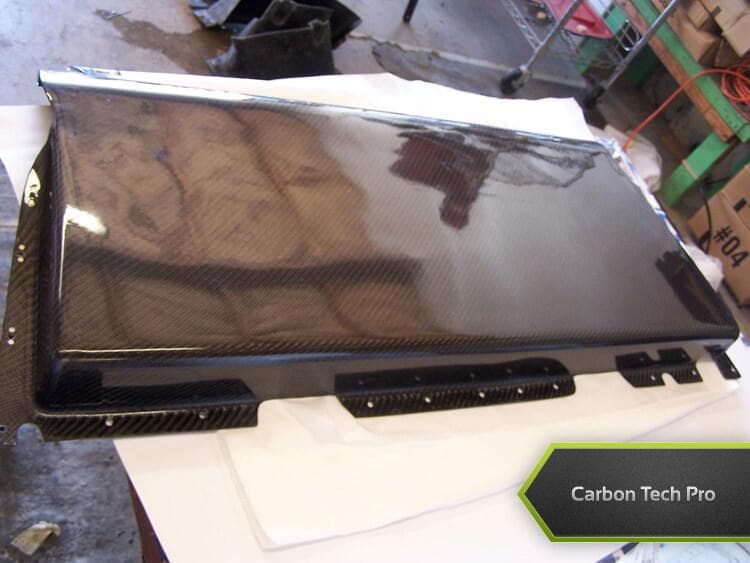 High-gloss woven carbon fiber panel fresh off the production line. High-gloss carbon fiber panel laying flat on a white padded surface in a workshop.